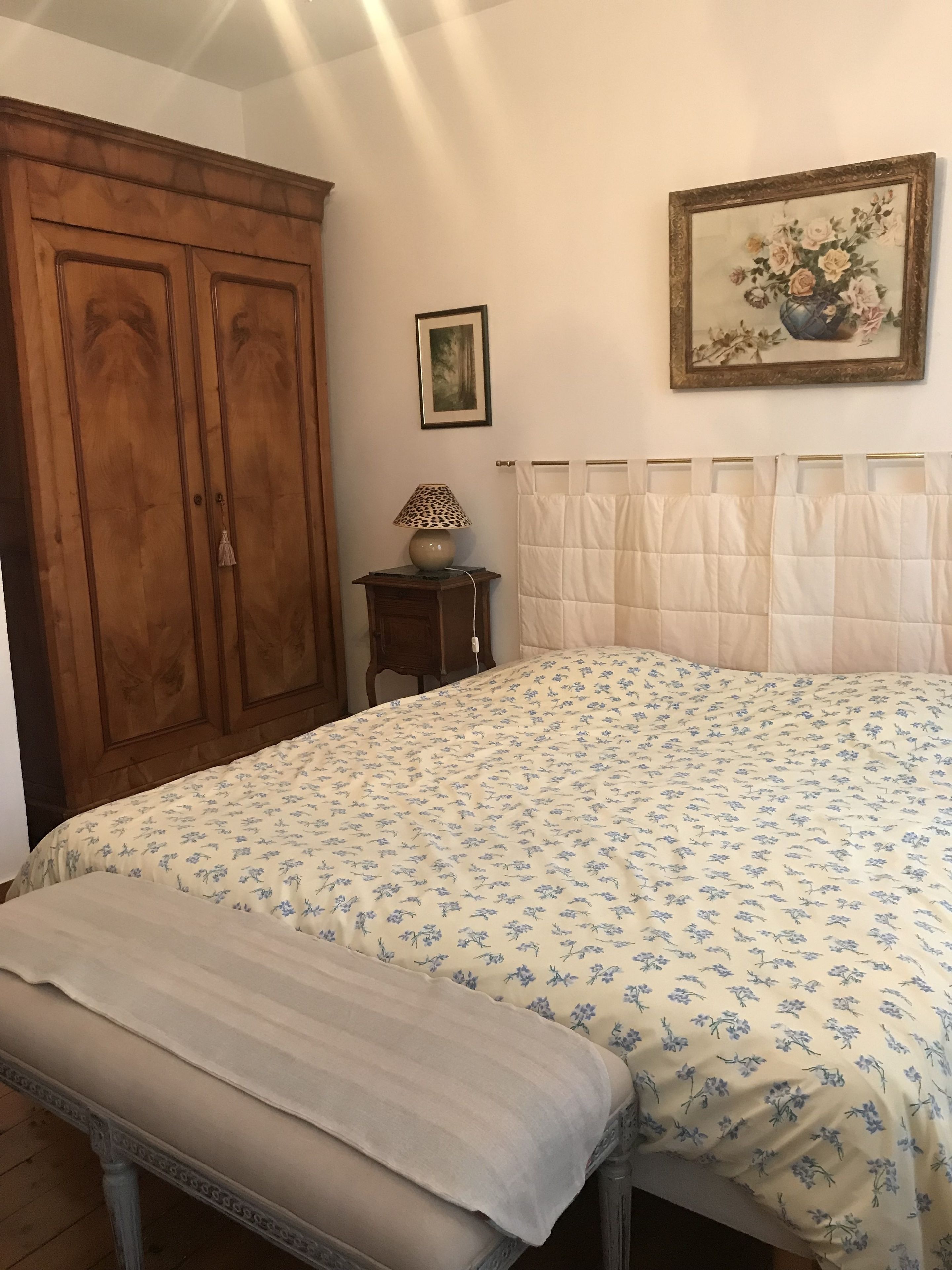 Un dormitorio con una cama grande, un armario de madera, una mesita de noche pequeña con una lámpara, un cuadro enmarcado en la pared y una ventana con cortinas.
