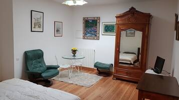 A small, well-lit room with a bed, a green chair, a small table with a vase, a mirror, and a radiator.