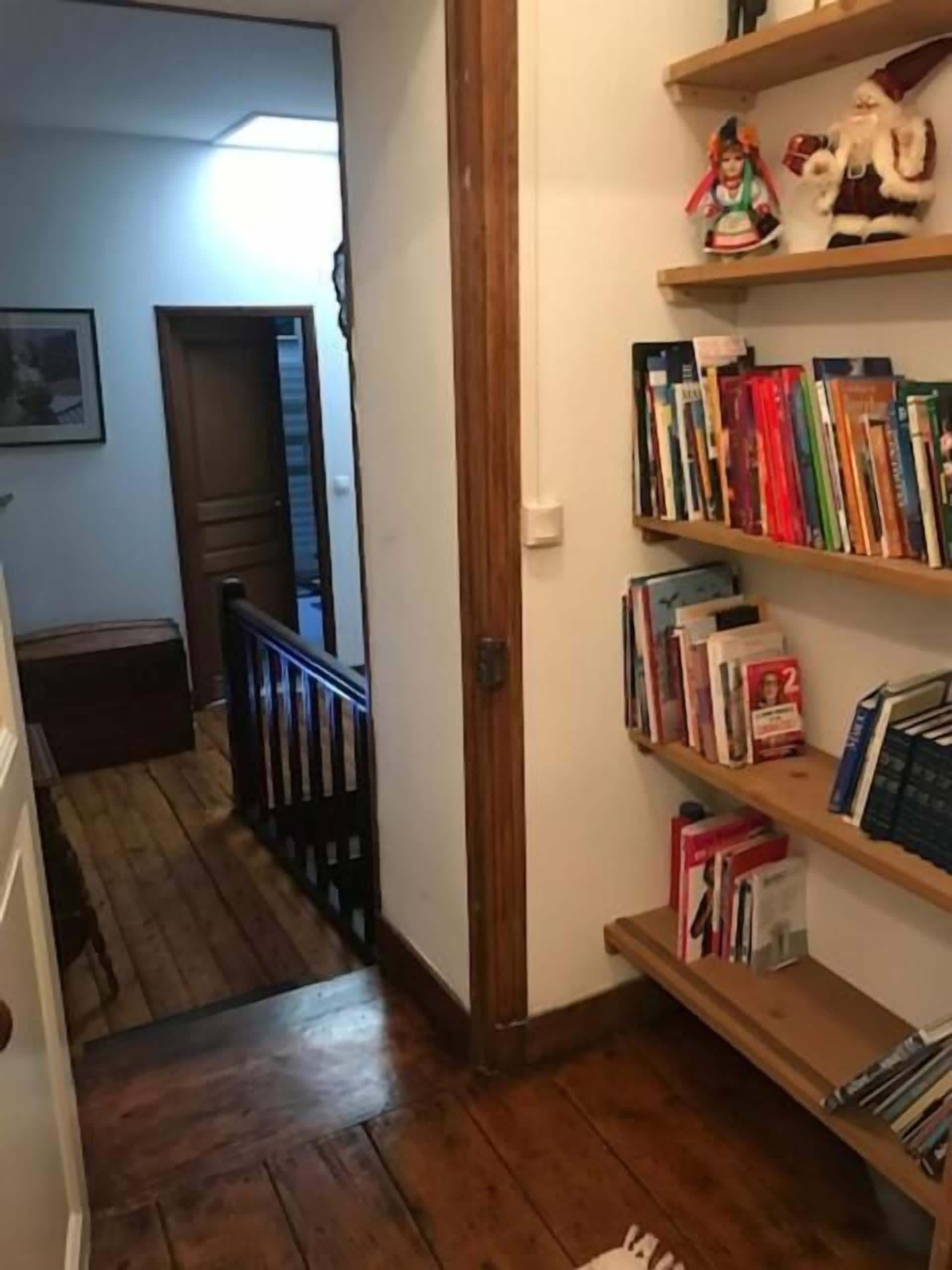 A hallway with wooden flooring, a staircase with a railing, and a bookshelf with various books and a Santa Claus figurine.