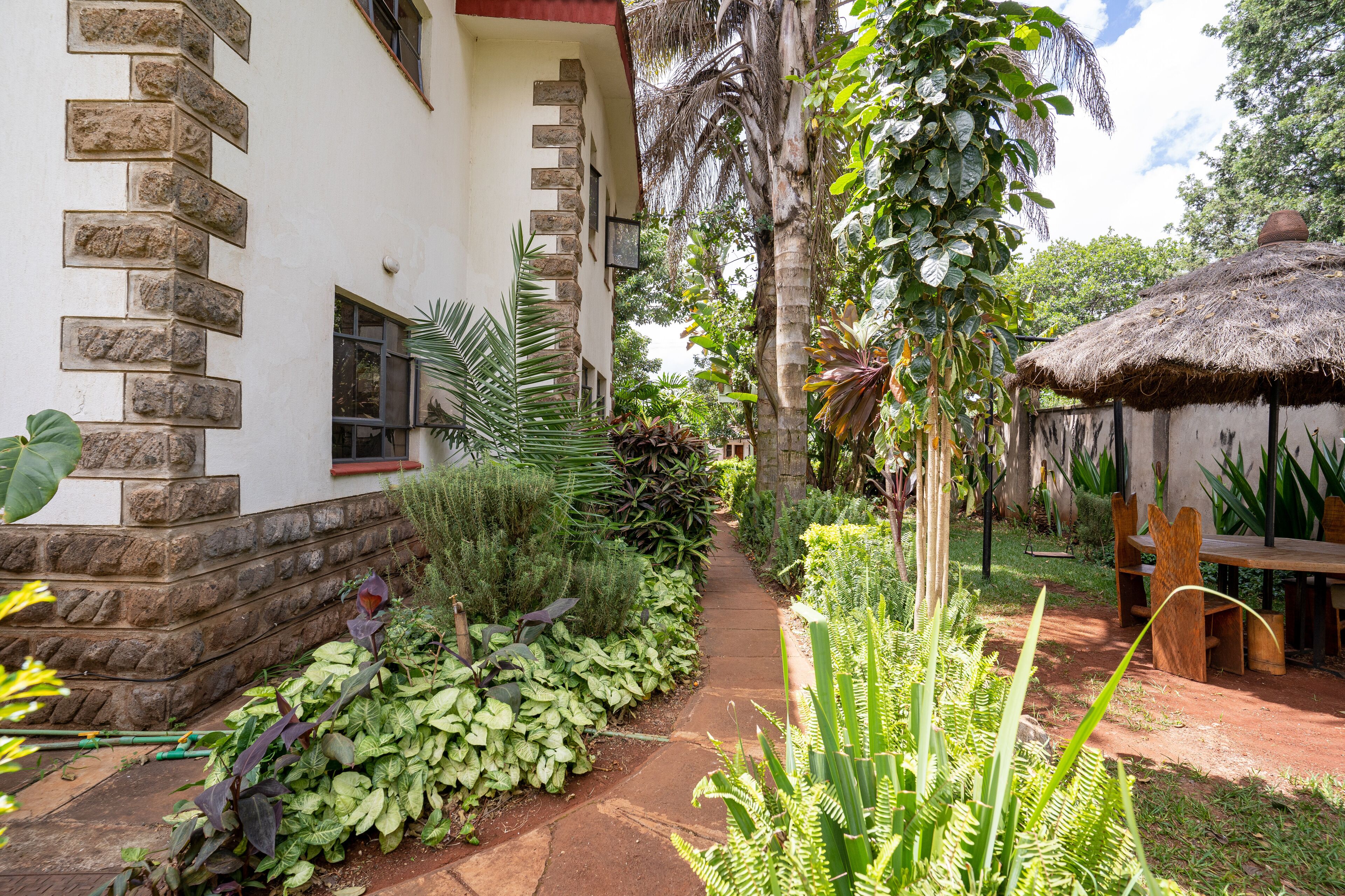 Uma trilha no jardim leva a uma construção com telhado de palha, com uma mesa e cadeiras de madeira.