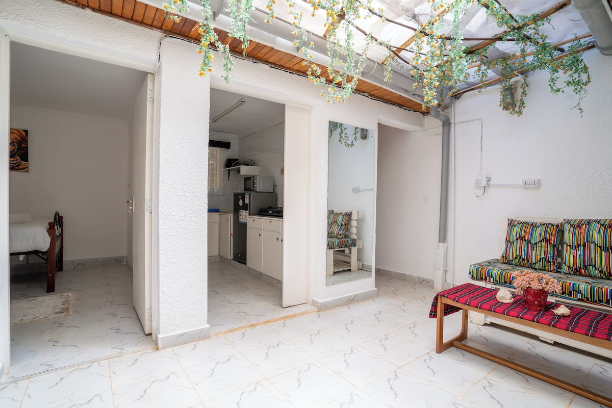 A room with a bed, a sofa, a dining table, and a kitchen area visible through an open doorway.