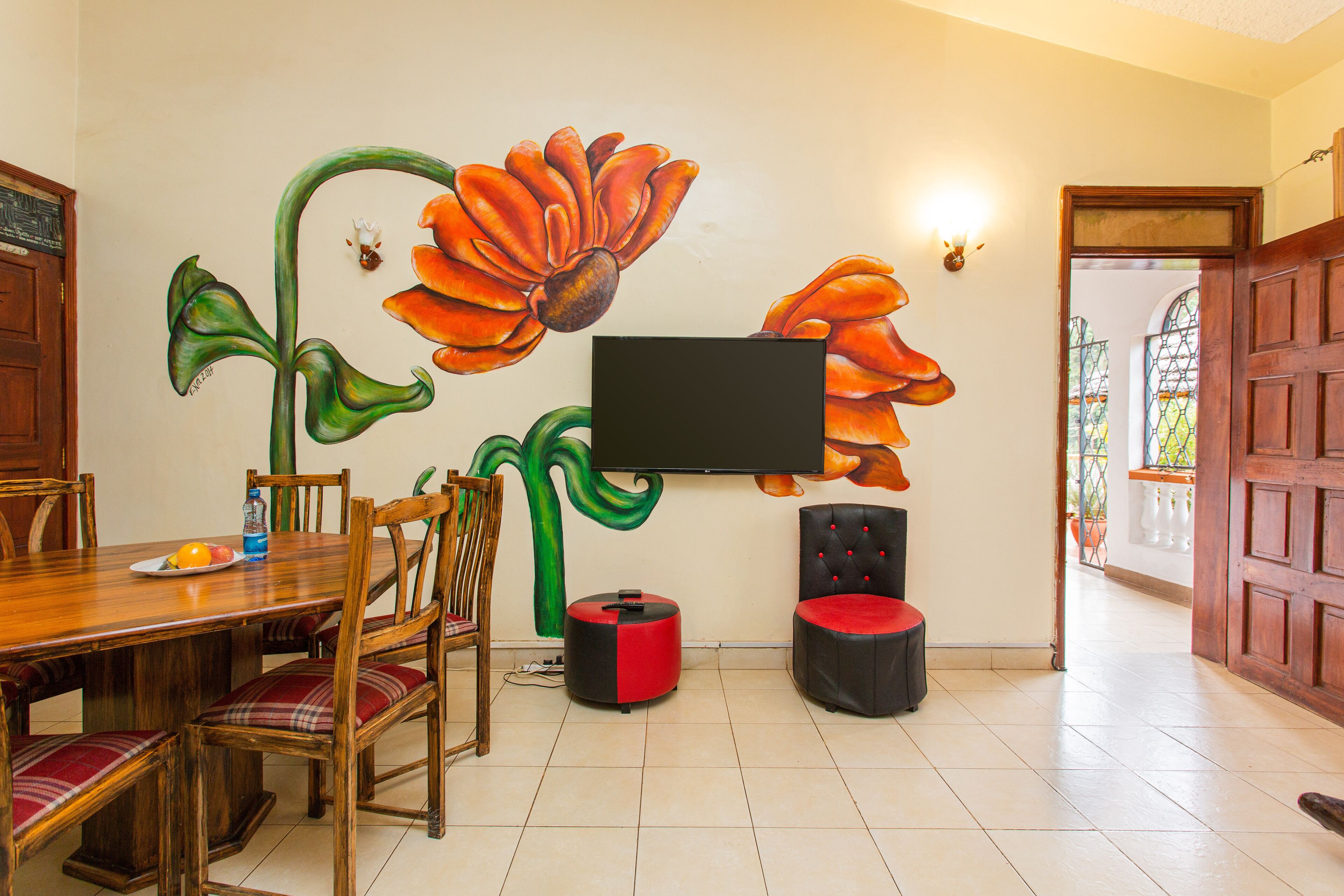 Un área de comedor con una mesa y sillas de madera, un televisor montado en la pared y un gran mural floral.