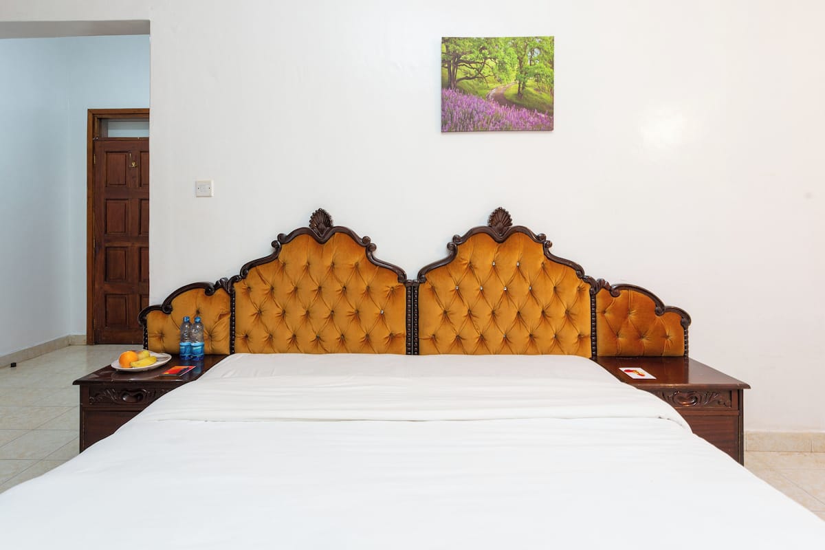 A bedroom with a wooden headboard, a bed, a nightstand with a plate of fruit and water bottles, and a painting on the wall.