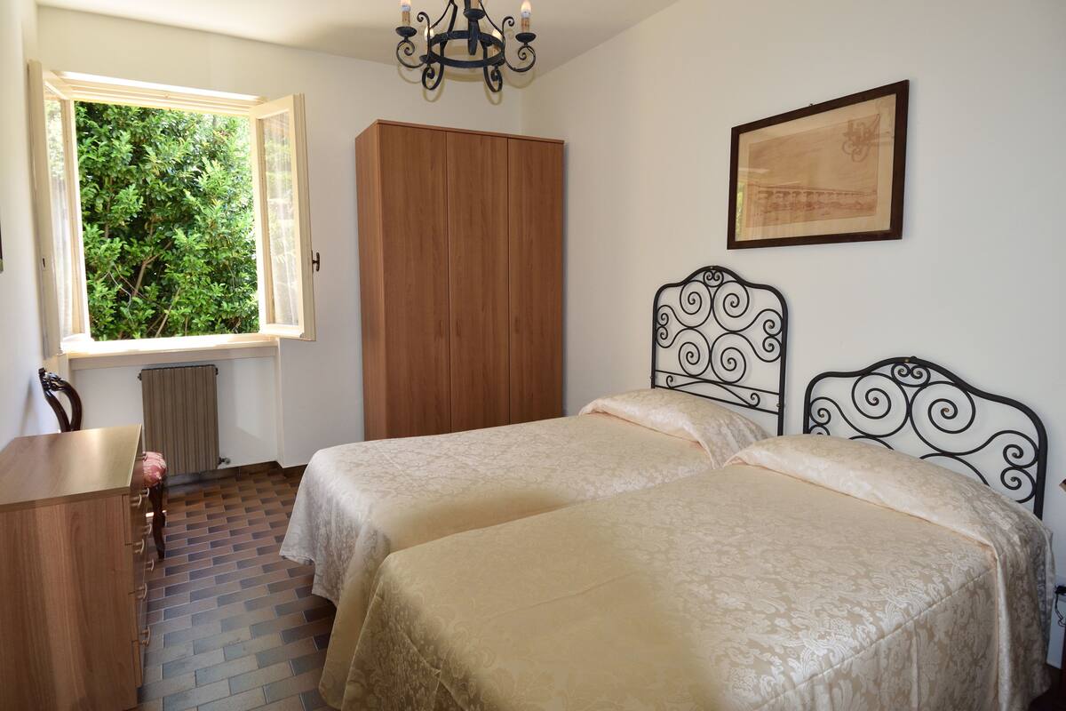 A bedroom with two beds, a wooden wardrobe, a desk, and a window with a view of greenery.