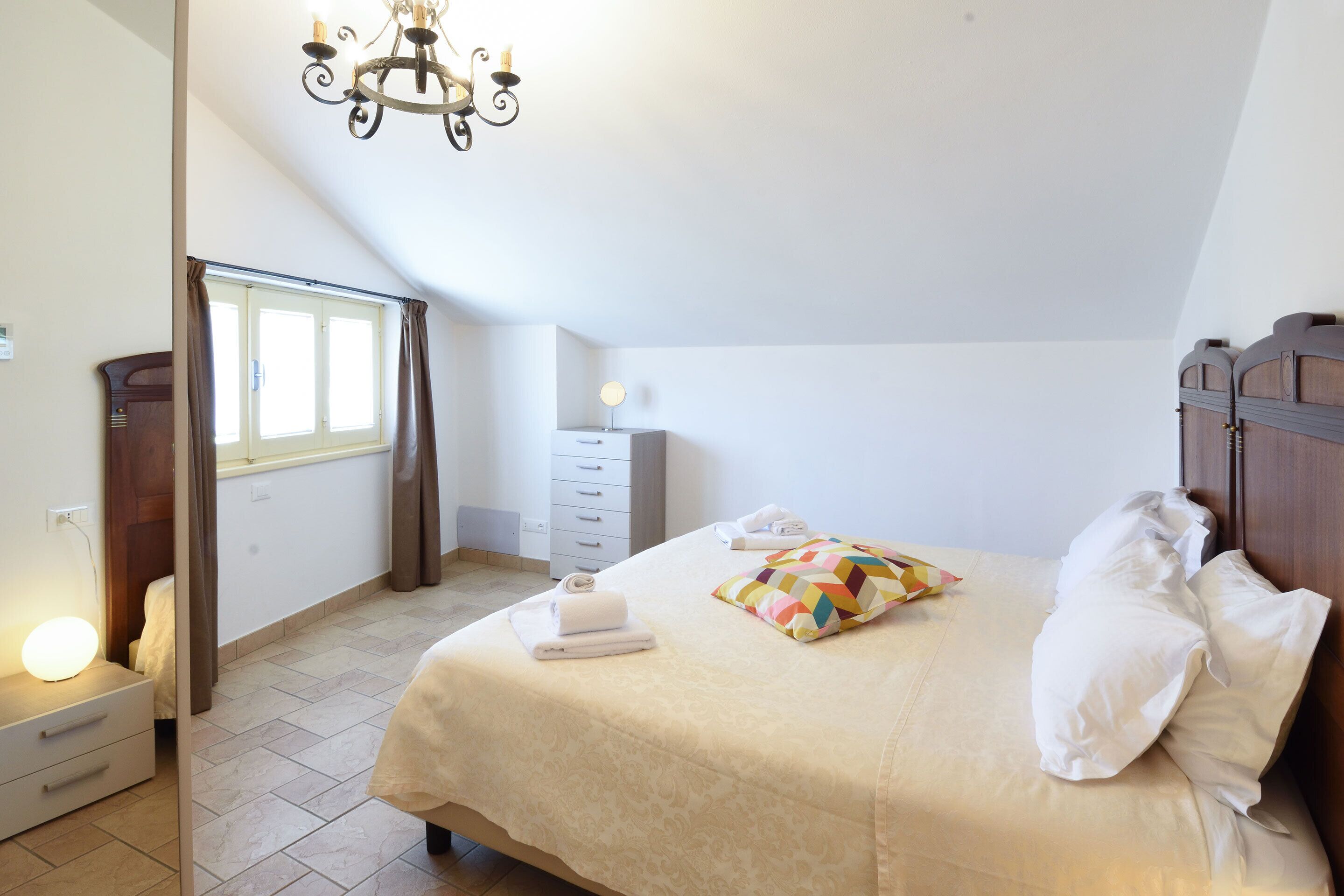 Ein Schlafzimmer mit Bett, Nachttisch, Kommode, einem Fenster mit Vorhängen und einer Chandelier.