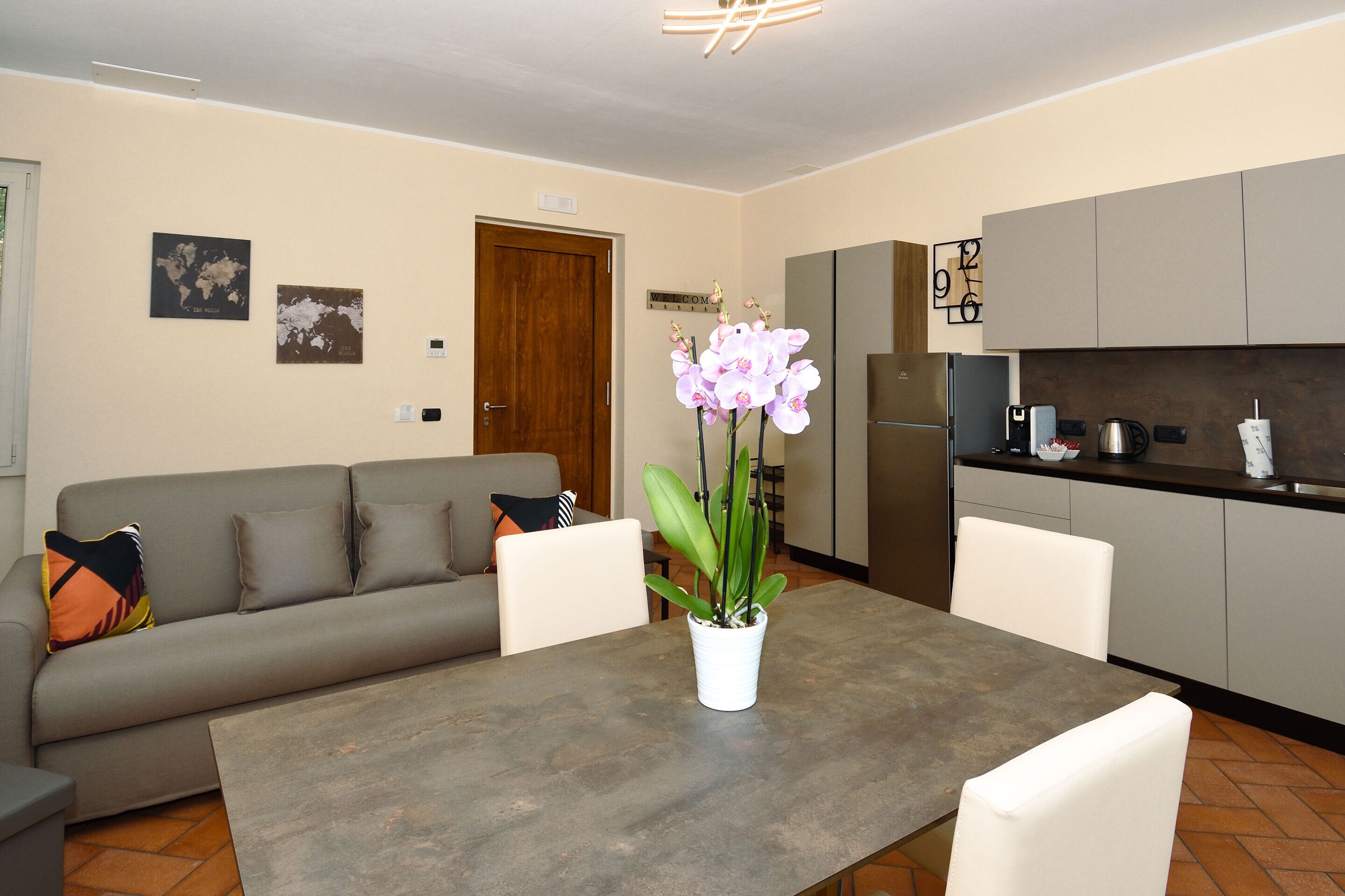 A modern living room with a grey sofa, a dining table with chairs, a potted orchid, and a kitchen area with cabinets and appliances.