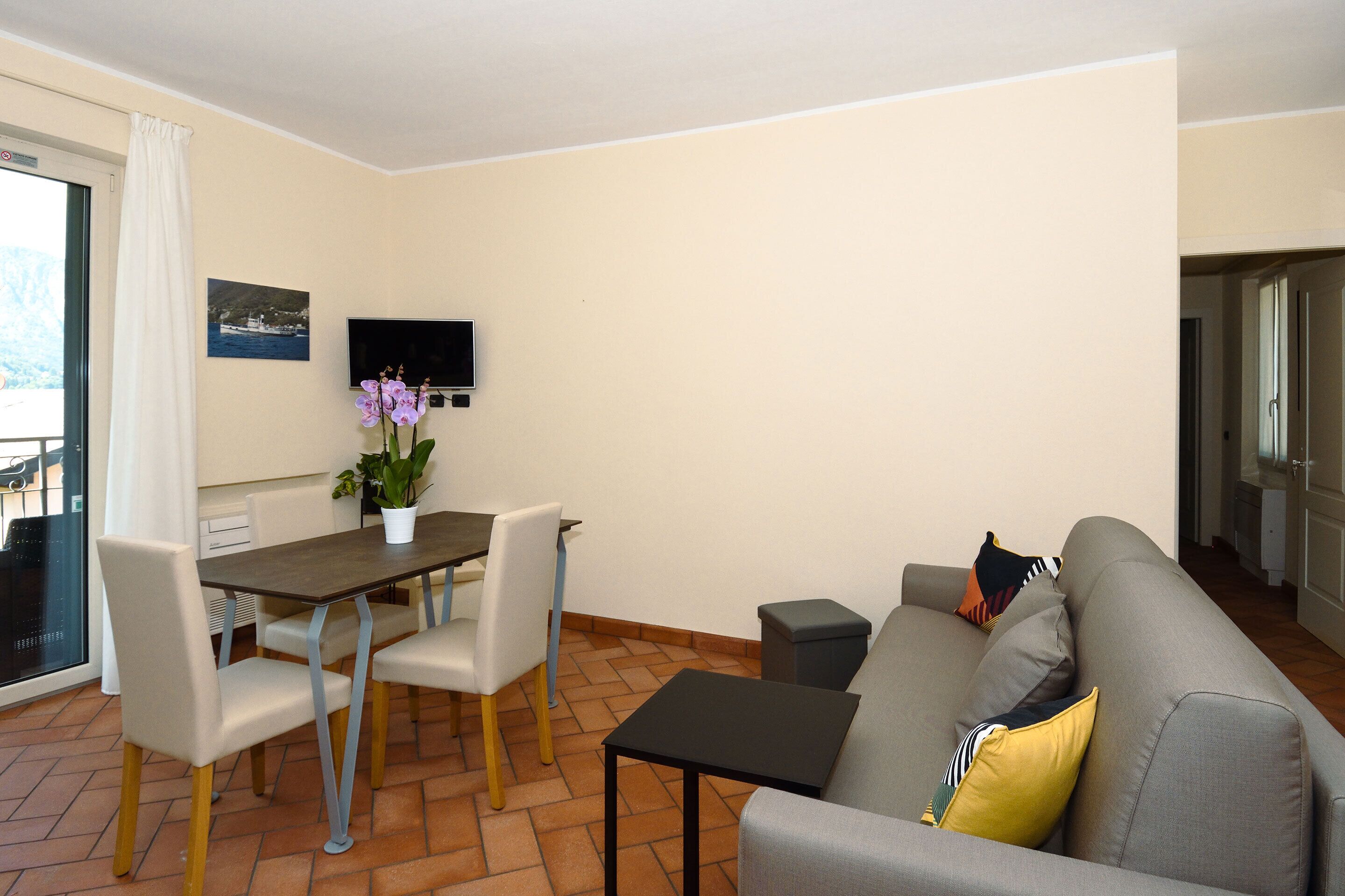 A living room with a sofa, a coffee table, a dining table with chairs, a potted plant, and a flat-screen TV mounted on the wall.