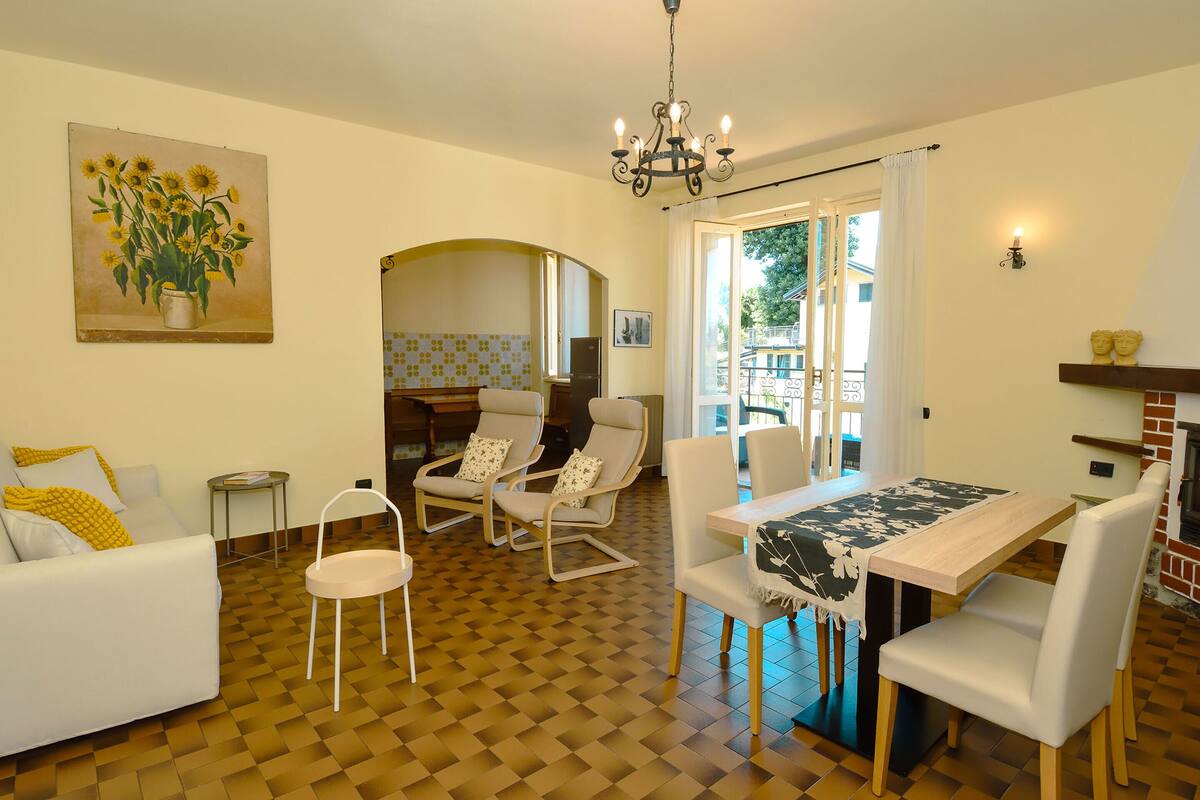 A dining area with a wooden table and chairs, a sofa with cushions, a painting of sunflowers, and a chandelier.