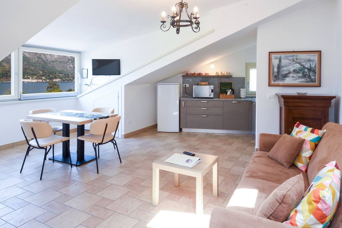 A modern living space with a dining area, a sofa, a small table, a kitchen, and a large window offering a view of the outdoors.