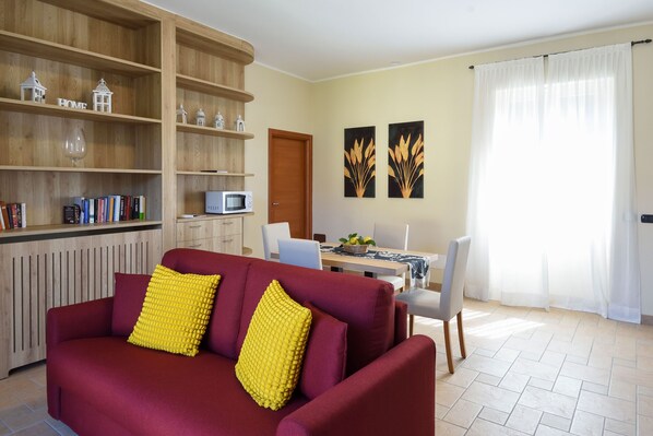 Un salon avec un canapé rouge, des coussins jaunes, une bibliothèque en bois, un coin repas avec une table et des chaises, et des œuvres d’art accrochées au mur.