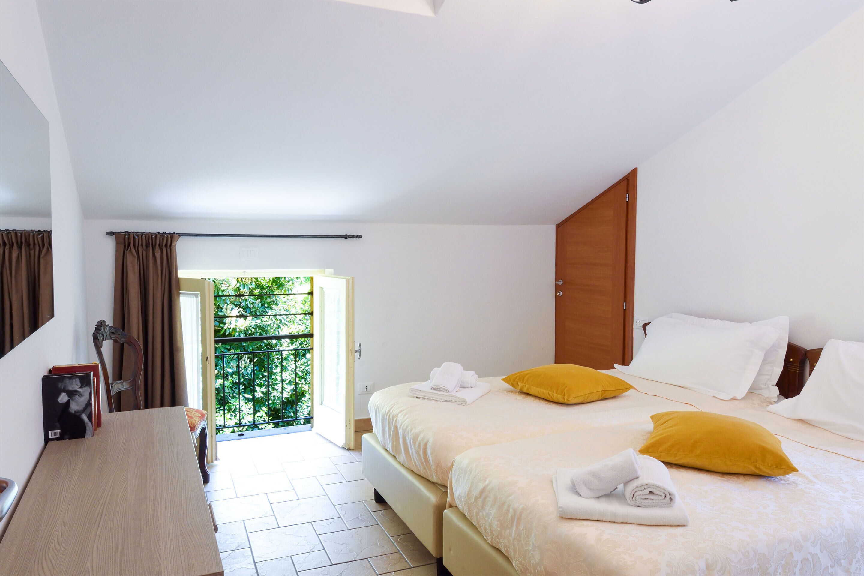 A hotel room with two beds, a wooden wardrobe, a desk, and a view of greenery through an open door.