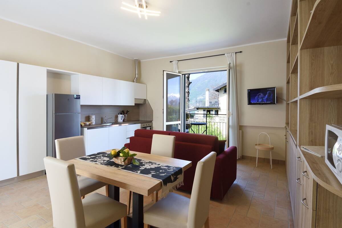 A compact kitchen with a dining area, a TV mounted on the wall, and a view of a mountain through a sliding door.