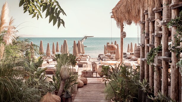 A beachside resort with a wooden deck leading to a lounge area, surrounded by thatched umbrellas and lounge chairs. - More Meni City Beach Rhodes (Rhodes)