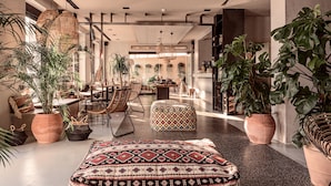 A spacious, modern living area with a large, patterned ottoman, wicker furniture, and abundant indoor plants. - More Meni City Beach Rhodes (Rhodes)