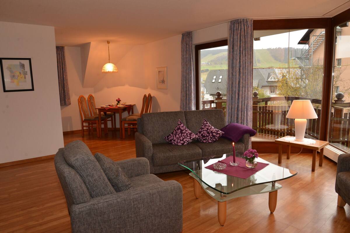 A living room with a sofa, armchair, glass coffee table, and a dining area with chairs and a table.