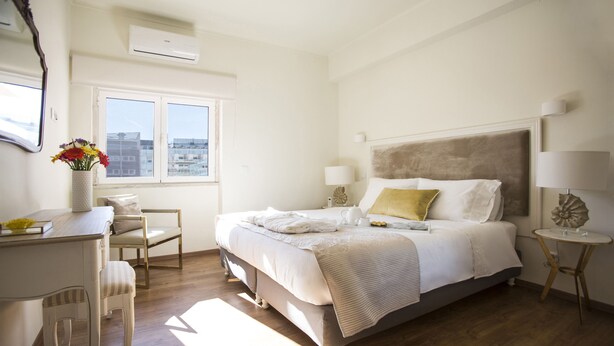 A hotel room with a bed, a desk, a chair, a window with a view of buildings, and a decorative lamp. - QUAD 3 Apartment with Balcony, Air Conditioned, Garage, City view, 24/7 security (Lisbon)