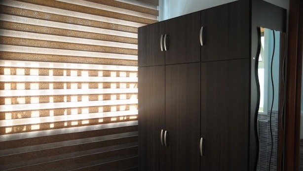 A room with a dark wooden wardrobe, a window with horizontal blinds, and a visible tiled floor.