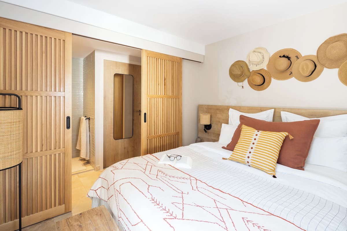 A bedroom with a bed, pillows, a bedside table, a wall-mounted hat display, and a wooden wardrobe.