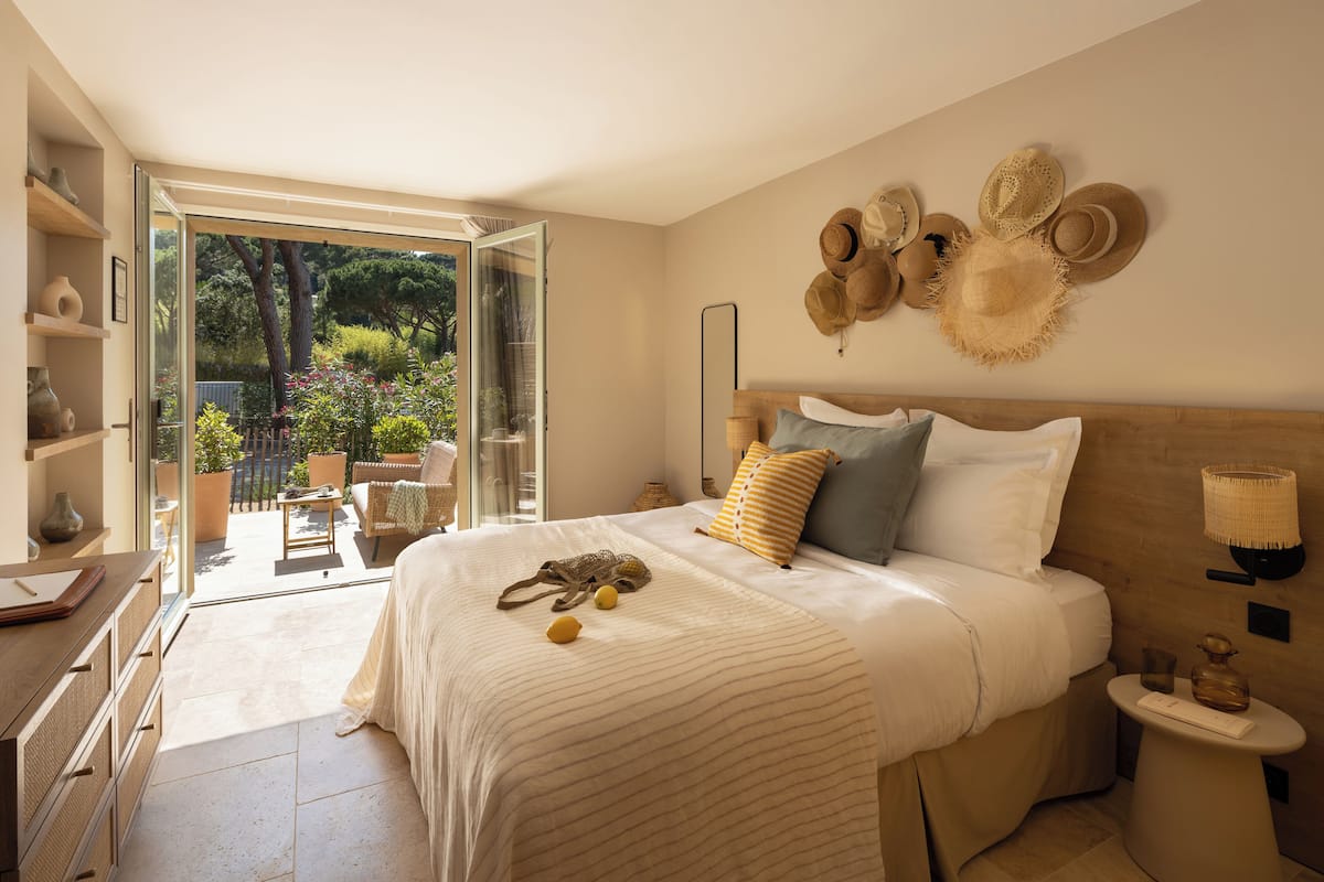 A bedroom with a large bed, a bedside table, a wall-mounted lamp, and a view of the garden through an open door.