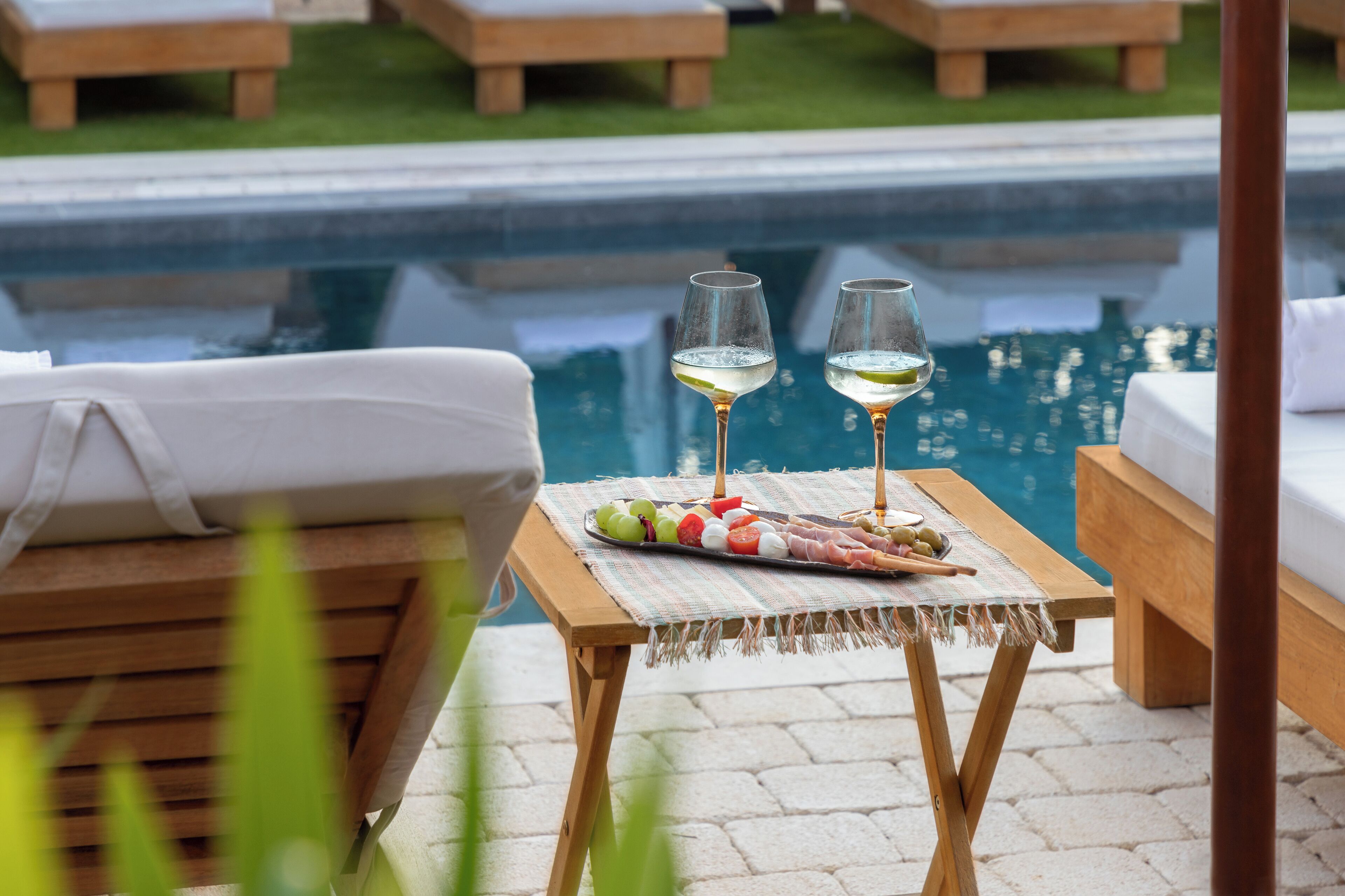 Ein Poolbereich mit Tisch für zwei Personen, mit einem Glas Weißwein, einer Platte mit verschiedenen Früchten und einer kleinen Schale Oliven.