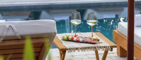 Ein Poolbereich mit Tisch für zwei Personen, mit einem Glas Weißwein, einer Platte mit verschiedenen Früchten und einer kleinen Schale Oliven.