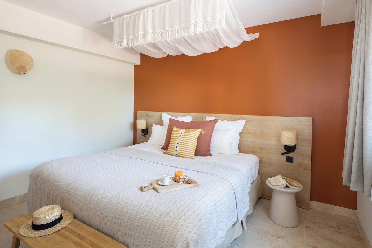 A neatly made bed with a striped blanket, a wooden nightstand with a hat, and a bedside table with a lamp and a tray of breakfast items.