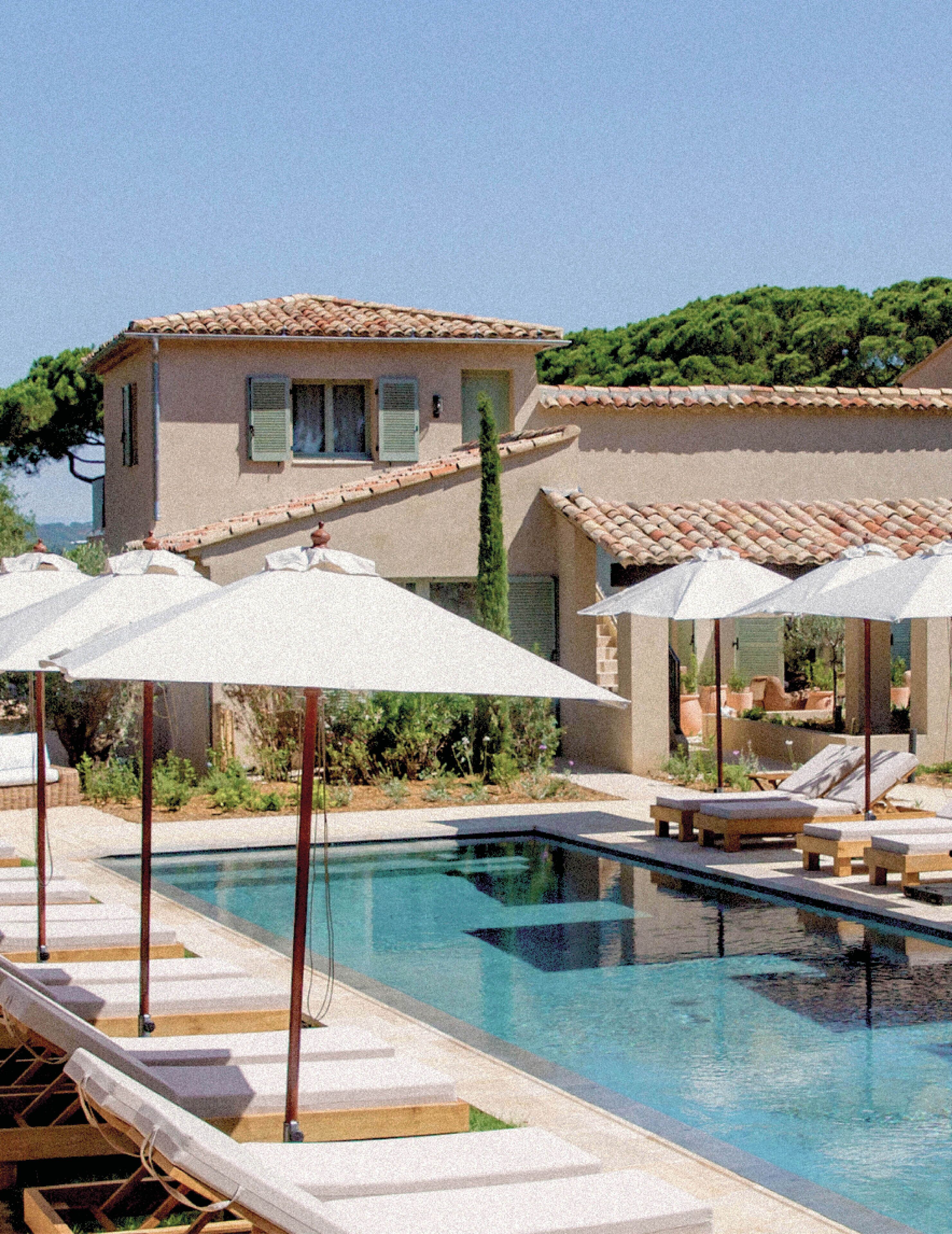 A swimming pool with lounge chairs and umbrellas, surrounded by a building with a tiled roof and a garden.