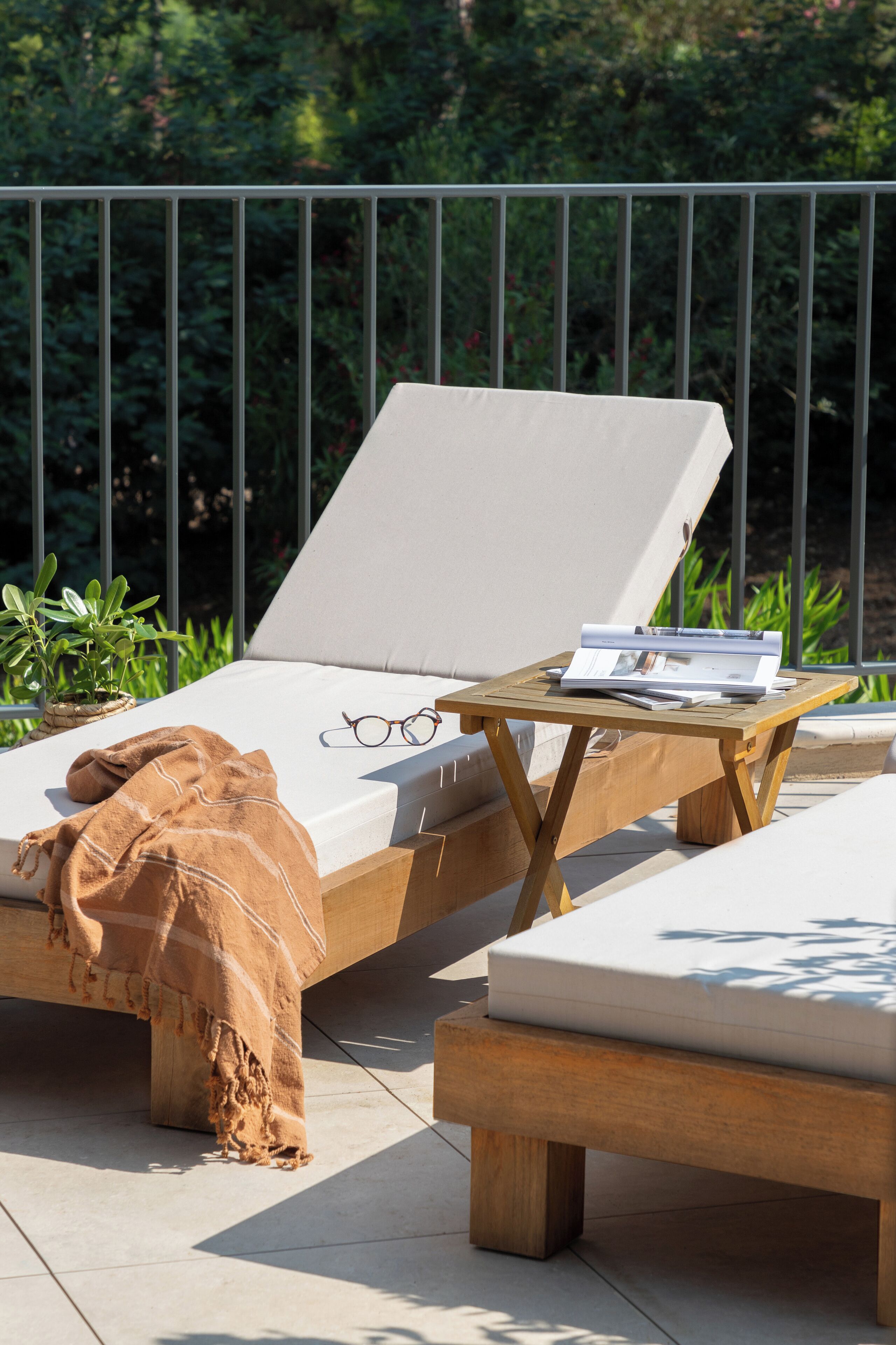 A sunlit outdoor lounge area with a cushioned seating area, a wooden table with a magazine and glasses, and a blanket on the floor.