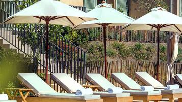 A poolside area with lounge chairs and umbrellas, a tiled roof, and a building with green shutters in the background.