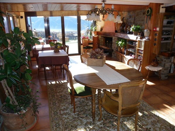 Un comedor acogedor con una mesa y sillas de madera, rodeado de plantas en macetas y vistas a montañas a través de amplios ventanales.