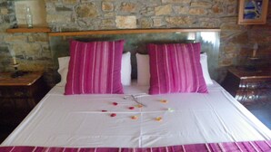 Una cama con ropa blanca y almohadas a rayas rosas, un muro de piedra al fondo y una pequeña estantería con botellas.
