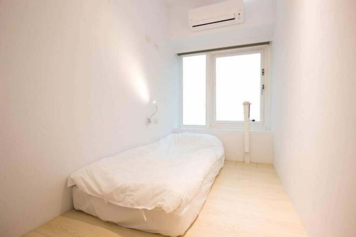 A small, single-bed room with a window, white walls, and a ceiling-mounted air conditioner.