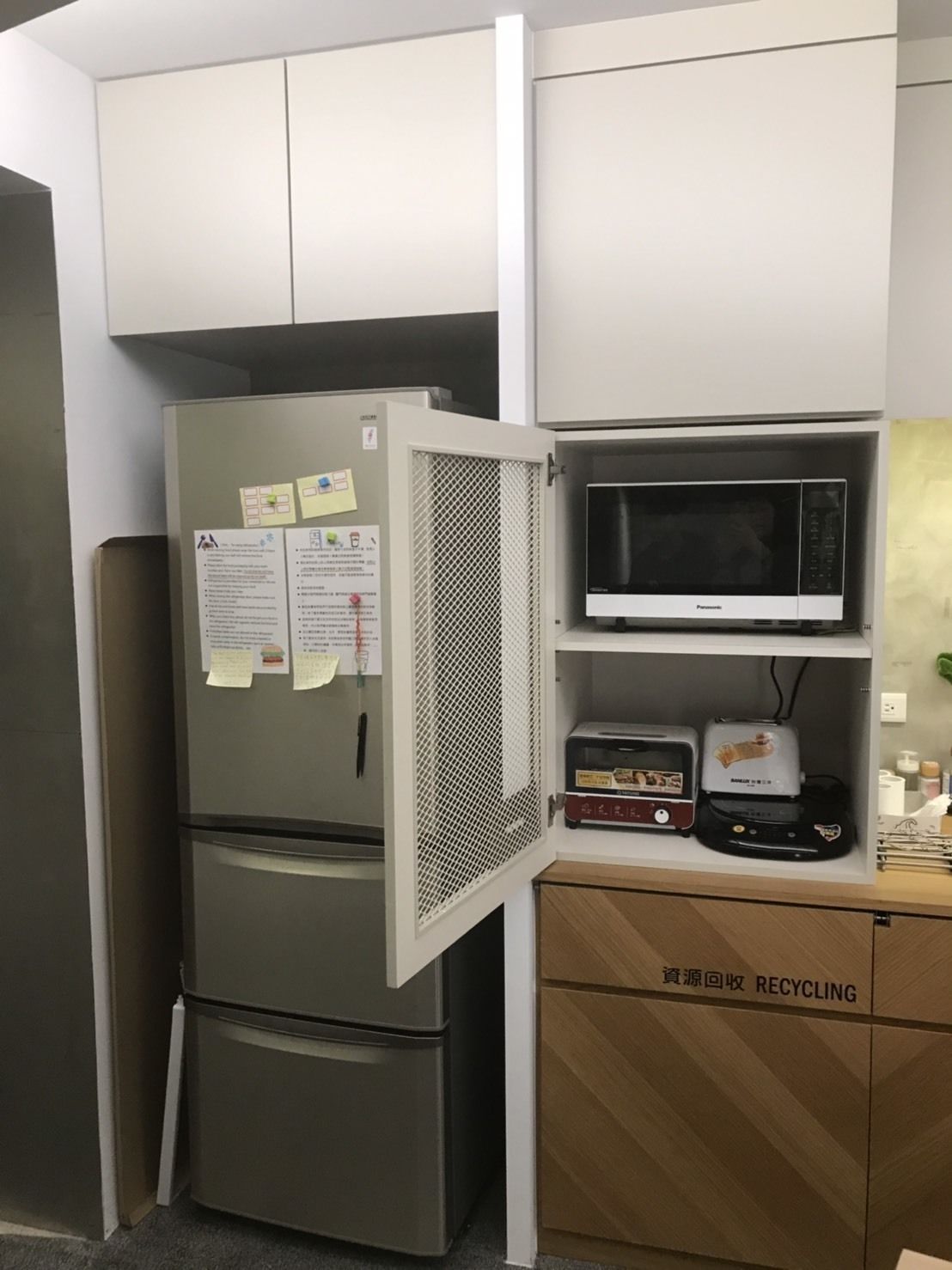 Cocina compacta con refrigerador de acero inoxidable, microondas y tostadora. Hay un contenedor para reciclaje con etiqueta en un idioma extranjero.