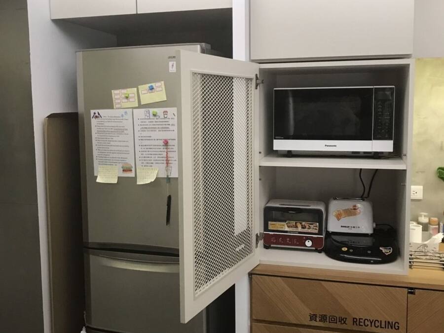 Cocina compacta con refrigerador de acero inoxidable, microondas y tostadora. Hay un contenedor para reciclaje con etiqueta en un idioma extranjero.