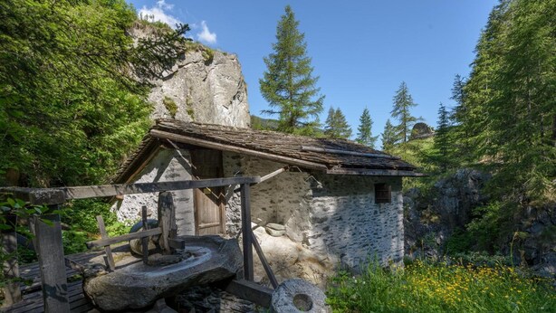 Ein rustikales Steinhaus mit Holzdach, umgeben von üppiger Vegetation und einer felsigen Klippe.