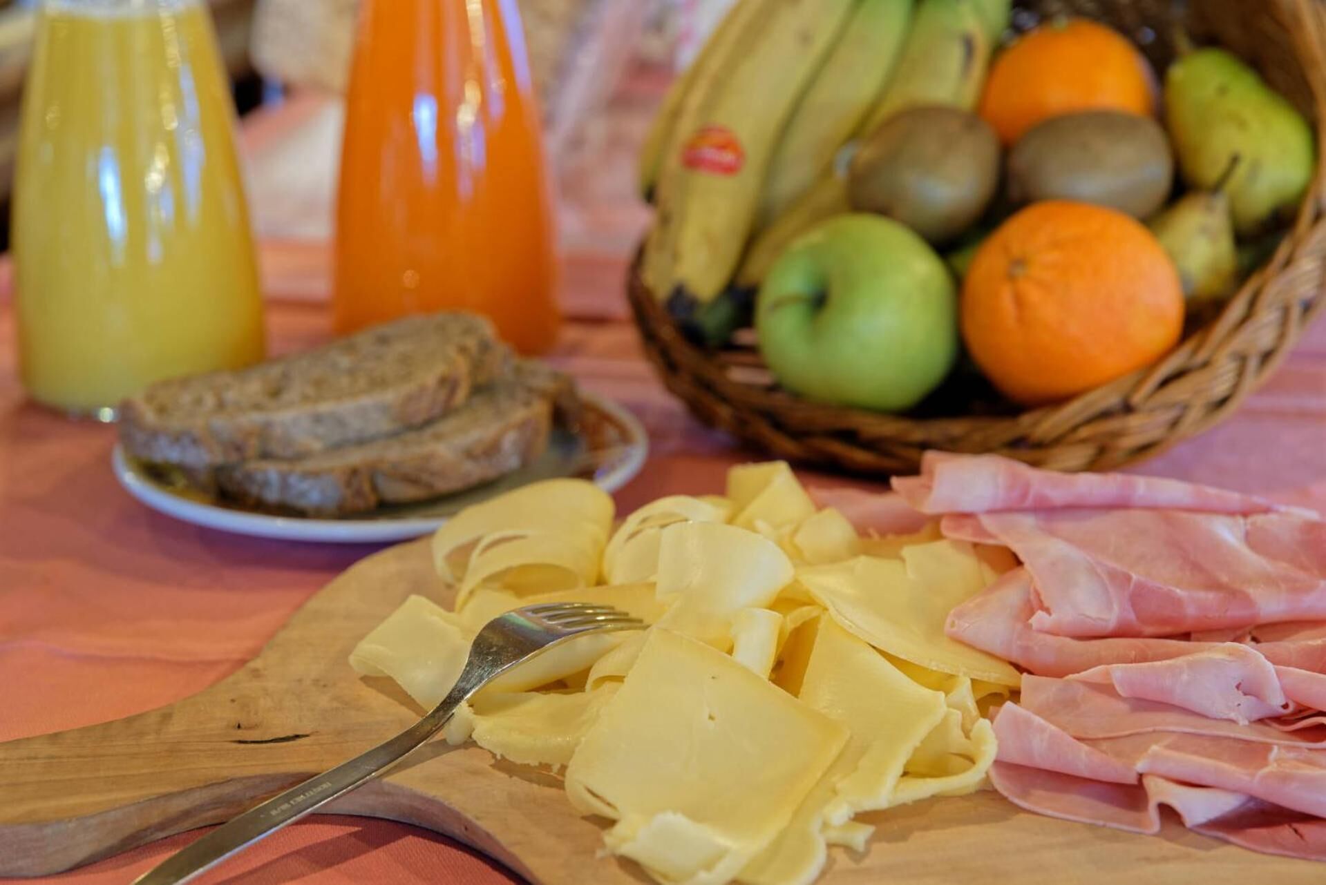 A breakfast table with sliced cheese, ham, bread, a fruit basket, and a bottle of orange juice.