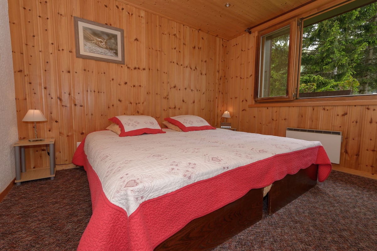 Une chambre à coucher aux murs lambrissés, avec deux lits, un tableau encadré et une fenêtre donnant sur des arbres.