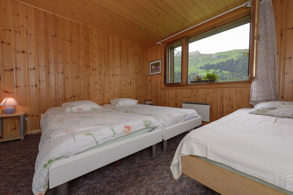 Une pièce aux murs lambrissés, avec deux lits, une fenêtre donnant sur des collines verdoyantes et une petite table de chevet avec une lampe.
