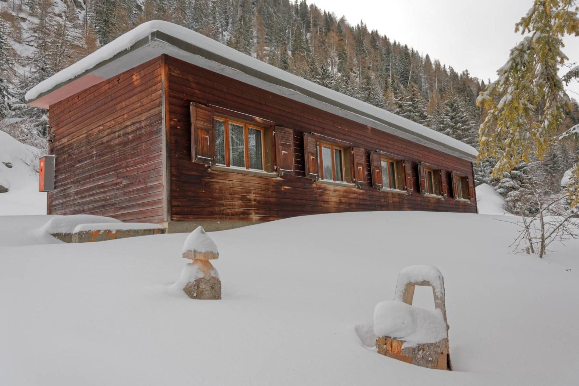 A wooden cabin with a sloped roof, surrounded by snow-covered trees and a mountainous backdrop.