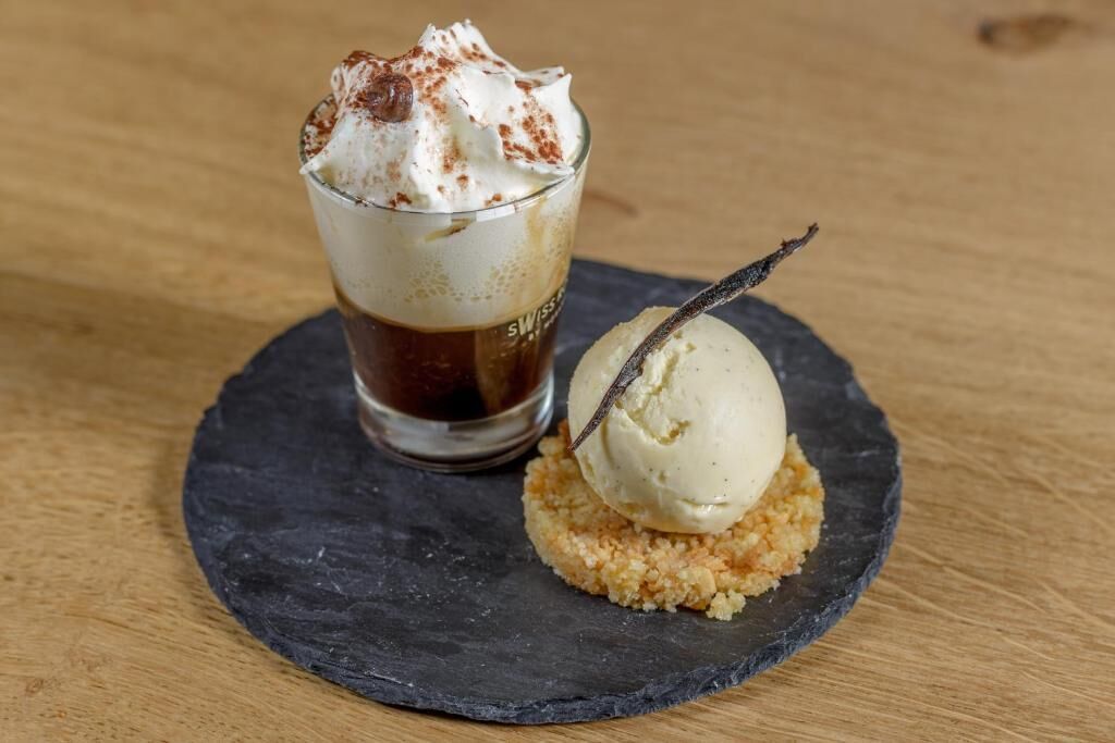Un postre que incluye un vaso de líquido oscuro, posiblemente café, cubierto de crema batida y una pizca de canela, junto con una bola de helado de vainilla sobre una base desmenuzable.