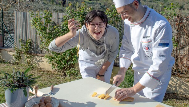 Two people preparing food outdoors with a scenic view of a historic town in the background. - B&B Il Fienile (San Gimignano)