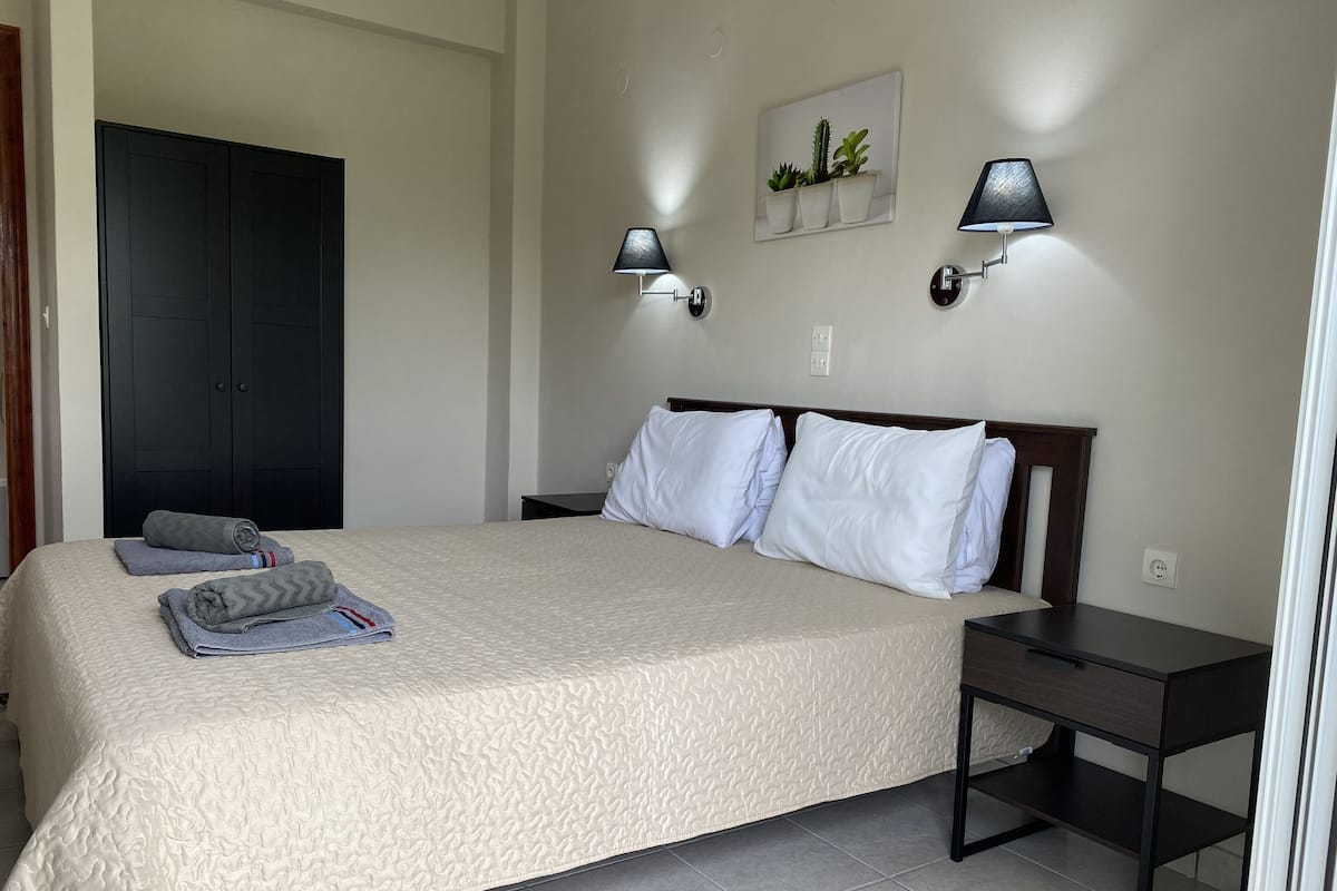 A neatly made bed with pillows and towels, a bedside table, a wall-mounted lamp, and a door in the background.