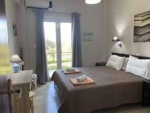 A hotel room with a large bed, a bedside table with a lamp, a view through the window, and a wall-mounted artwork. - Alexander Apartments (Corfu)