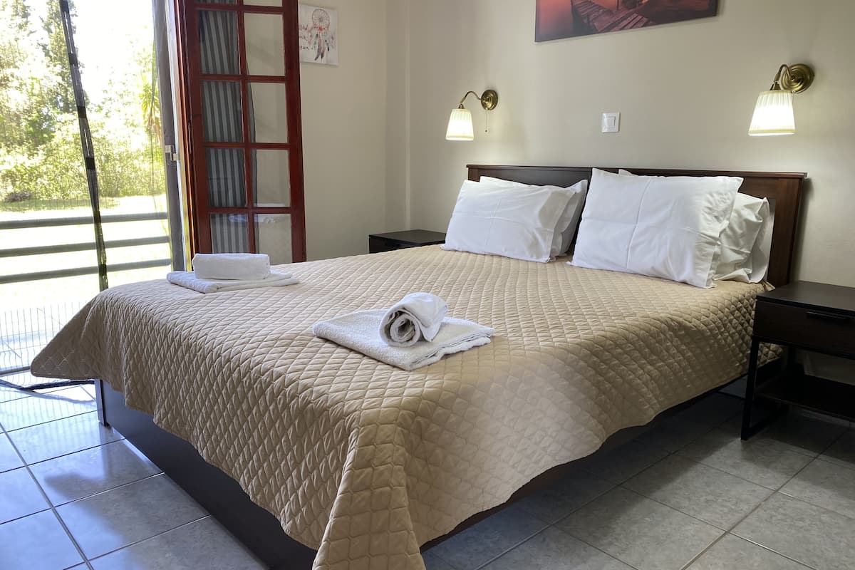A neatly made bed with a quilted beige bedspread, white pillows, and rolled white towels on the side. There are two bedside tables with lamps, a wall-mounted artwork, and a view of greenery outside.