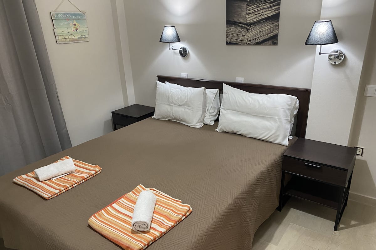 A neatly made bed with two towels, a headboard with three pillows, two bedside tables, and two wall-mounted lamps.