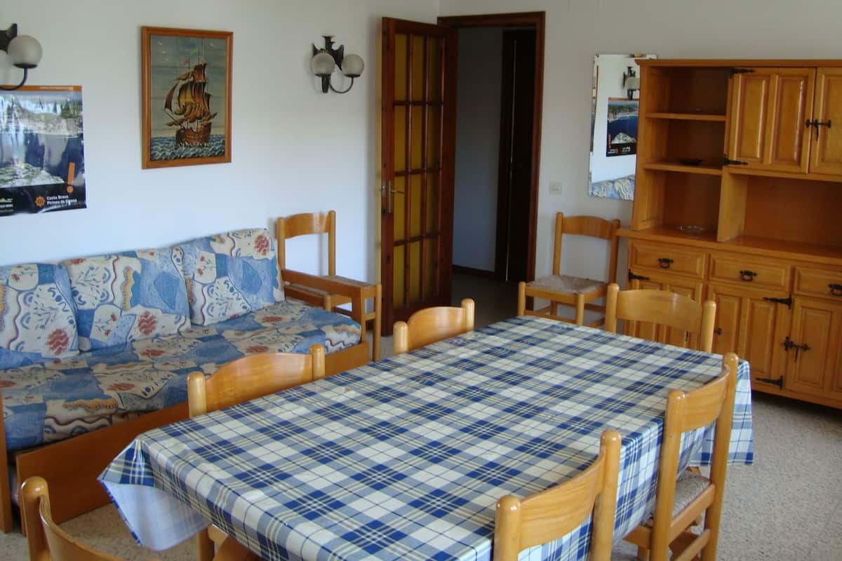 A dining area with a checkered tablecloth, wooden chairs, a sofa with a floral pattern, and wooden cabinets.