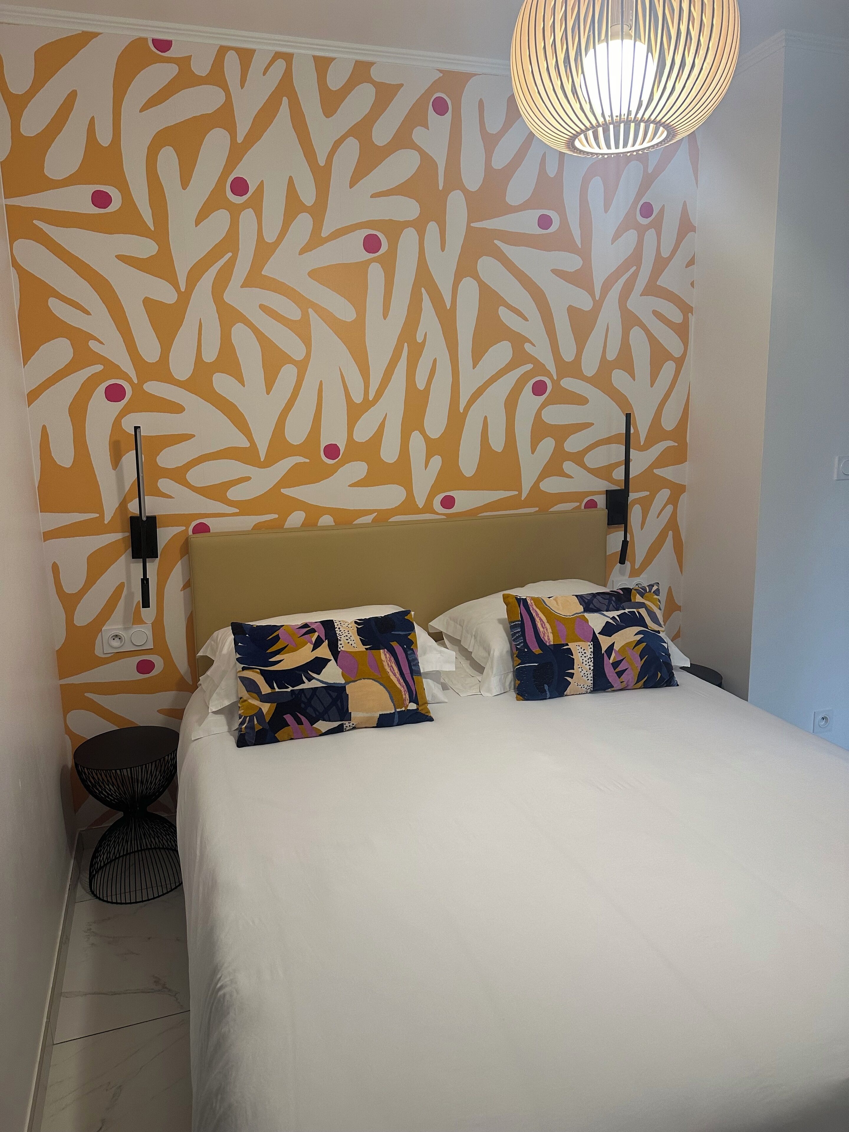 Ein Schlafzimmer mit einem Bett, zwei Kissen, einer Wand mit Blättermotiv, einer Pendelleuchte und einem kleinen runden Tisch.