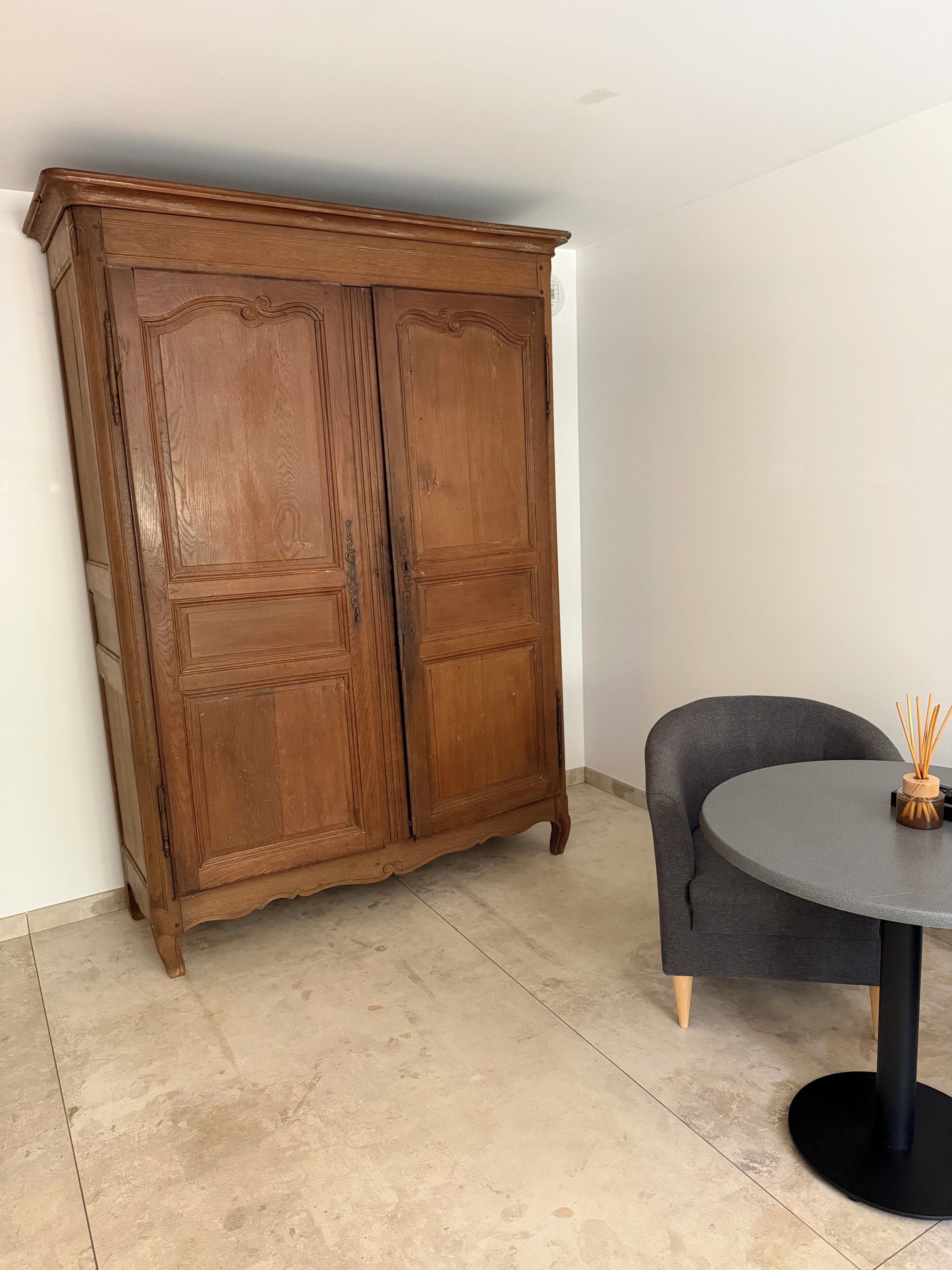 Una habitación con un armario de madera, una mesa redonda con una silla oscura y un florero con ramas de juncos.