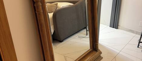 A large mirror with ornate wooden frame, a bed with grey bedding, and a floor with white marble tiles.