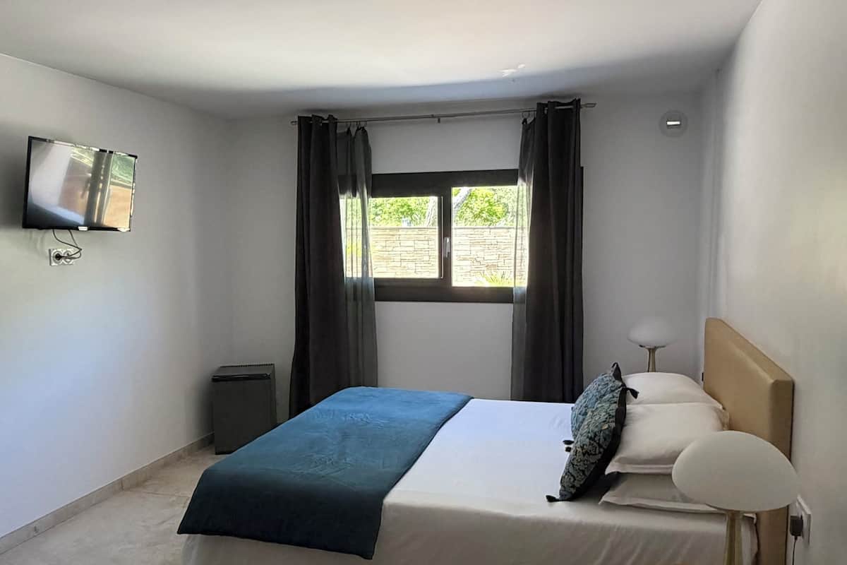A bedroom with a bed, a bedside table with a lamp, a TV mounted on the wall, and a window with curtains.